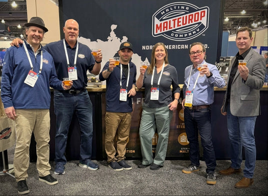 Six people standing together at a trade show booth, holding up glasses of malt beverage in front of a large Malteurop Company backdrop. The group is wearing branded lanyards, and the setting is a convention hall with booths and displays visible in the background.